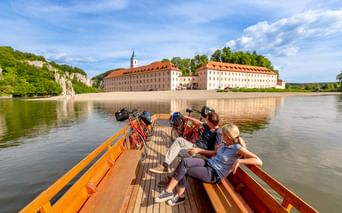 Radfahrer auf Holzfähre überqueren die Donau zum Kloster Weltenburg. Das barocke Kloster mit orangen Dächern liegt an Kalksteinfelsen.