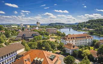 Aerial view of Hameln with red-tiled roofs, the Weser River flowing through, a bridge, and green hills under a blue sky with white clouds.