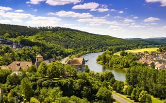 Luftaufnahme der Burg Homburg mit Turm neben einem Fluss, umgeben von bewaldeten Hügeln, grünen Wiesen und Dorfhäusern unter blauem Himmel.