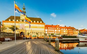 Emder Rathaus mit gelb-weißer Fassade und Turm hinter einer hölzernen Hafenpromenade mit Flaggen. Orangefarbene Backsteingebäude und Boote im Hafen.