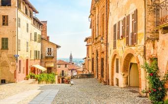 Kopfsteinpflasterstraße in Saluzzo mit ockerfarbenen historischen Gebäuden, Fensterläden und einem Kirchturm in der Ferne.