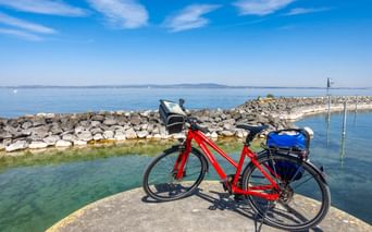 Rotes Tourenrad mit blauen Taschen auf steinerner Mole am Bodensee. Steinerner Wellenbrecher und ruhiges blaues Wasser unter klarem Himmel.
