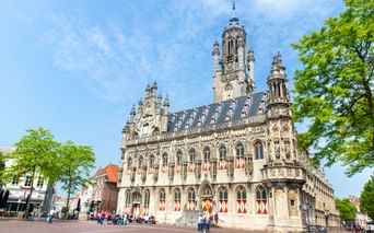 Gotisches Rathaus von Middelburg mit verzierter Fassade, hohem Turm und rot-weißen Fensterläden. Besucher auf dem Platz unter blauem Himmel.