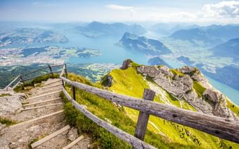 Bergweg mit Holzzaun auf dem Pilatus mit Blick auf den Vierwaldstättersee. Grüne Almwiesen, Felsgipfel und blauer See mit umliegenden Bergen.