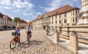 Zwei Radfahrer auf gepflastertem Platz in Eichstätt mit historischen Barockgebäuden und verziertem Säulenmonument unter blauem Himmel.