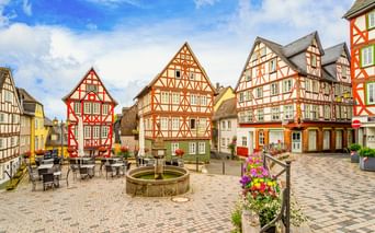 Domplatz square in Wetzlar with colorful half-timbered houses, a stone fountain, outdoor café seating, and flower-decorated benches under blue sky.