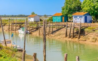 Kanal mit Holzpfählen und kleinem Boot, gesäumt von bunten Hütten mit orangen Dächern in Marennes, Frankreich, unter blauem Himmel.