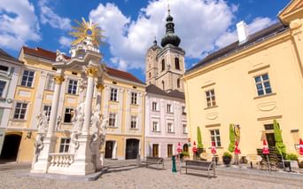 Baroque square in Krems-Stein with ornate white monument, pastel buildings, and church tower under blue sky with white clouds.