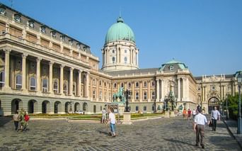 Burgpalast in Budapest mit seiner markanten grünen Kuppel und gelber Barockfassade. Besucher gehen über den Kopfsteinpflaster-Innenhof.