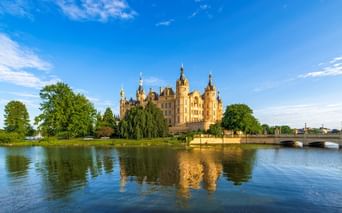 Schweriner Schloss, ein prächtiger Palast mit goldenen Kuppeln und Türmen, liegt auf einer Insel umgeben von ruhigem Seewasser unter blauem Himmel.