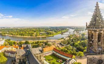 Luftaufnahme von Magdeburg mit gotischem Domturm im Vordergrund, Elbe schlängelt sich durch grüne Landschaft, Stadtgebäude unter blauem Himmel.