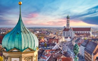 Luftaufnahme von Augsburg mit grünen Zwiebeltürmen im Vordergrund und dem hohen Rathausturm über der historischen Altstadt bei Sonnenuntergang.