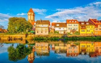Spitzer Turm, ein runder Turm mit orangefarbenem Dach in Wertheim, spiegelt sich im Main. Bunte Fachwerkhäuser säumen das Flussufer.