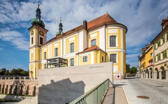 Gelb-weiße Barockkirche mit grünen Kuppeltürmen in Donaueschingen. Steinmonument mit Aufschrift 'Donauquelle' im Vordergrund.