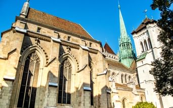 St. Peter's Cathedral in Geneva with Gothic arched windows, stone facade, and distinctive green copper spire against a clear blue sky.