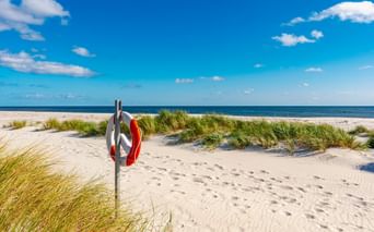 Weißer Sandstrand in Dueodde auf Bornholm mit rot-weißem Rettungsring an Stange, Dünen mit Strandgras und blauem Meer unter klarem Himmel.