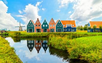 Reihe bunter traditioneller Häuser mit Treppengiebeln, die sich in einem Kanal spiegeln. Grüne Wiesen umgeben das Wasser, eine Windmühle ist in der Ferne sichtbar.