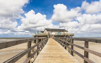 Langer Holzsteg führt zu einem Pfahlbau am Strand von St. Peter-Ording. Breiter Sandstrand und dramatischer Wolkenhimmel im Hintergrund.