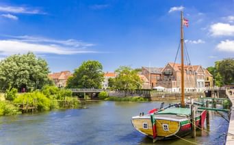 Gelb-grünes traditionelles Boot im Hafen von Ribe mit bunten historischen Gebäuden und einer Brücke über den Fluss unter blauem Himmel.