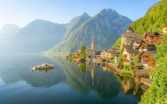 Hallstätter See mit traditionellem Alpendorf am Ufer, Kirchturm, Bergen und einem Boot auf ruhigem Wasser, das die Szenerie spiegelt.