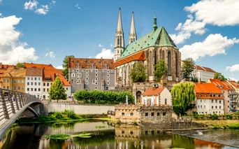 St. Peter and Paul Church with twin spires and green copper roof in Görlitz, reflected in the Neisse River with historic buildings and bridge.