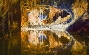 Beleuchtete Feengrotte Saalfeld mit farbigen Stalaktiten und Stalagmiten, die sich in stillem unterirdischem Wasser spiegeln, mit goldener und blauer Beleuchtung.