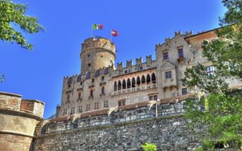 Schloss Buonconsiglio in Trient mit Steinmauern, zinnenbesetzten Türmen und italienischen Flaggen vor blauem Himmel, eingerahmt von grünen Bäumen.