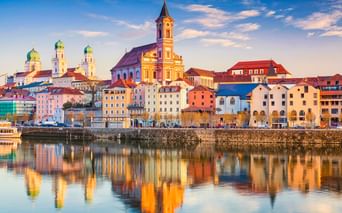 Passau old town skyline with colorful buildings along the Danube riverfront. Church towers with green domes and a yellow church are reflected in the water.