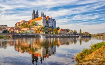 Historisches Meißen mit Albrechtsburg und Dom auf dem Burgberg über der Elbe. Bunte Häuser säumen das Ufer, perfekt im ruhigen Wasser gespiegelt.