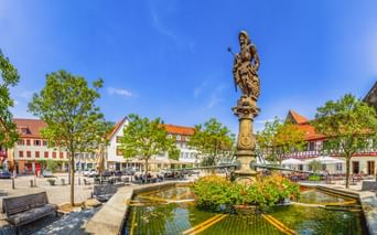 Historischer Brunnen mit Statue auf Säule auf dem Öhringer Marktplatz, umgeben von bunten Blumen, Bäumen, Bänken und traditionellen Gebäuden.