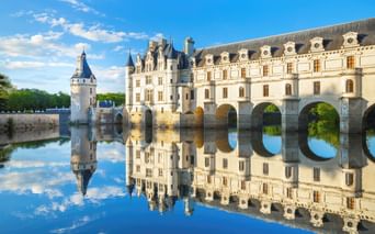 Schloss Chenonceau mit weißer Steinfassade und Bogenbrücke über ruhigem Wasser, das sich perfekt unter blauem Himmel spiegelt.