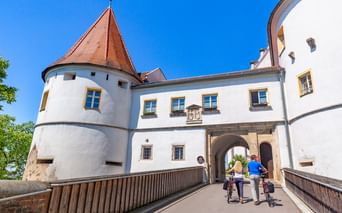 Two cyclists walking their bikes across a bridge toward Wörth Castle entrance. White castle walls with red-tiled tower under blue sky.