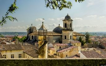 Blick auf eine historische Kirche mit Kuppel und Glockenturm in Beaucaire, umgeben von Steingebäuden mit Terrakottadächern unter blauem Himmel.