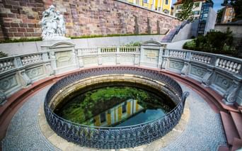 Rundes Steinbecken mit verziertem Eisengeländer markiert die Donauquelle in Donaueschingen, umgeben von weißen Balustraden und einer Statue.