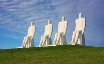 Vier monumentale weiße sitzende Figuren auf einem Grashügel vor blauem Himmel in Esbjerg. Die Skulpturen blicken zum Meer.