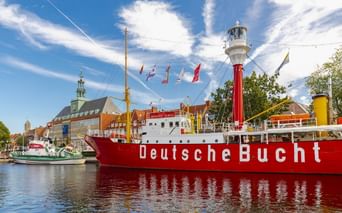 Rotes Feuerschiff Deutsche Bucht mit weißem Leuchtturm im Hafen von Emden. Historische Gebäude und blauer Himmel mit Wolken im Hintergrund.