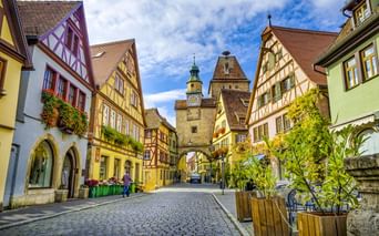 Kopfsteinpflasterstraße in Rothenburger Altstadt mit bunten Fachwerkhäusern und mittelalterlichem Torturm mit Uhr unter blauem Himmel.