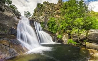 Wasserfall Cascada del Diablo in Villanueva de la Vera, der in ein natürliches Becken fließt, umgeben von üppiger grüner Vegetation und Felsen.