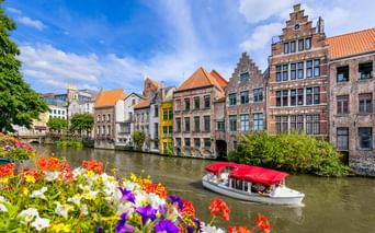 Ausflugsboot mit rotem Dach auf Kanal in Gent vor historischen Treppengiebelhäusern. Bunte Blumen rahmen die Uferszene unter blauem Himmel.