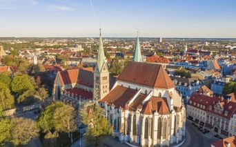 Luftaufnahme des Augsburger Doms mit zwei Türmen und roten Ziegeldächern, umgeben von historischen Gebäuden und Stadtlandschaft unter blauem Himmel.