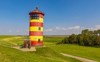 Rot-gelb gestreifter Pilsumer Leuchtturm in Krummhörn auf grüner Wiese mit geparkten Fahrrädern unter blauem Himmel.