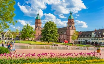 Freudenstädter Stadtkirche mit Zwillingstürmen und grünen Kupferdächern mit Blick auf einen Brunnenplatz mit bunten Tulpen und Menschen an einem sonnigen Tag.