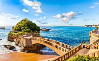 Steinerne Fußgängerbrücke mit Balustraden zu einer grünen Felseninsel in Biarritz. Blaues Meer, Sandstrand und Küstenlandschaft unter Sonnenhimmel.