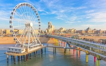 Scheveningen Strand mit weißem Riesenrad auf Pier und buntem Steg über dem Wasser. Sandstrand und Gebäude unter blauem Himmel sichtbar.