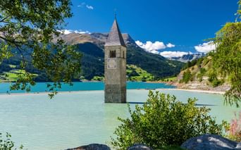 Historischer Kirchturm ragt aus dem türkisfarbenen Wasser des Reschensees mit Bergen im Hintergrund, eingerahmt von grünem Laub unter blauem Himmel.