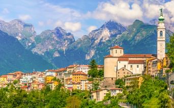 Historische Stadt Belluno mit bunten Gebäuden und einer Kirche mit Glockenturm vor dramatischen Berggipfeln unter blauem Himmel.