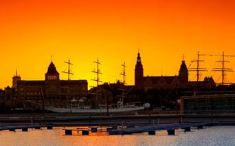 Silhouette der historischen Uferpromenade Stettins bei Sonnenuntergang mit Kirchtürmen und Segelschiffmasten vor orange-gelbem Himmel.