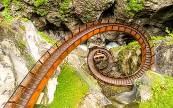 Luftaufnahme eines spiralförmigen Holzstegs in der Liechtensteinklamm mit moosbewachsenen Felswänden und fließendem Wasser.