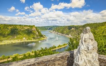 Steinerne Loreley-Statue in St. Goarshausen mit Blick auf den Rhein, der sich durch grüne Hügel schlängelt, mit einem Kreuzfahrtschiff auf dem Wasser.