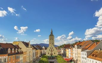 Historischer Stadtplatz in Deggendorf mit dem hohen Glockenturm des Alten Rathauses im Zentrum, umgeben von bunten Gebäuden und Bäumen.